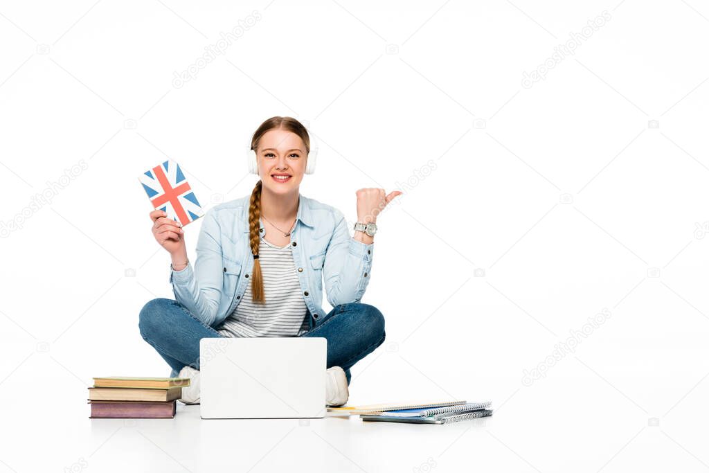 smiling girl learning English on floor in headphones near laptop, books and copybook and pointing with finger aside isolated on white