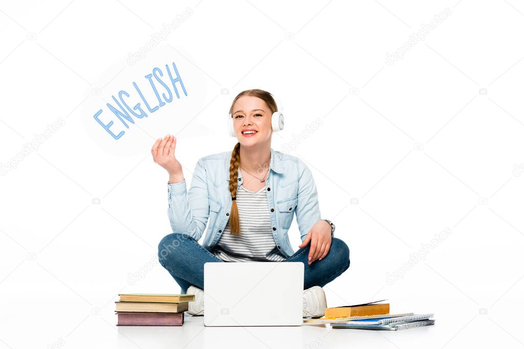 smiling girl sitting on floor in headphones with speech bubble with english lettering near laptop, books and copybooks isolated on white