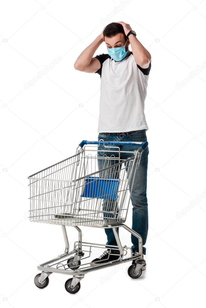 shocked man in medical mask looking at empty shopping cart isolated on white