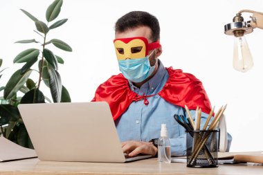 Tıbbi maskeli ve süper kahraman kostümlü bir adam beyaz dizüstü bilgisayar kullanıyor. 
