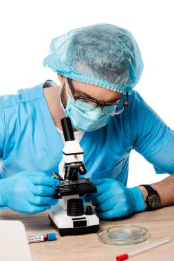 Tıbbi şapkalı ve gözlüklü bilim adamı test tüplerinin yanında mikroskoba dokunuyor ve beyaz üzerinde koronavirüs harfleri var. 