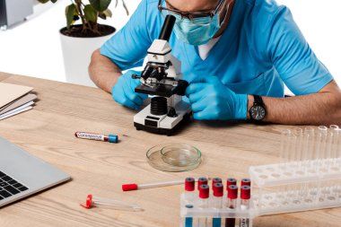 Tıbbi maskeli ve gözlüklü bilim adamı mikroskopla test tüplerinin yakınına bakıyor ve beyaz üzerinde koronavirüs harfleri var. 