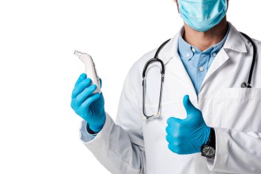 Tıbbi maskeli doktor ve beyaz önlüklü, temas edilmeyen ateş ölçer tutan ve beyaz üzerinde izole edilmiş baş parmağını gösteren... 
