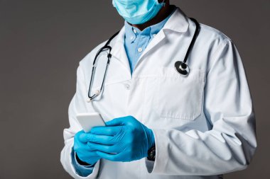 Tıbbi maskeli ve beyaz önlüklü doktorun gri üzerinde izole edilmiş akıllı telefonu kullanması.