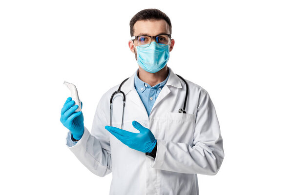 doctor in medical mask and white coat pointing with hand at non-contact pyrometer isolated on white 