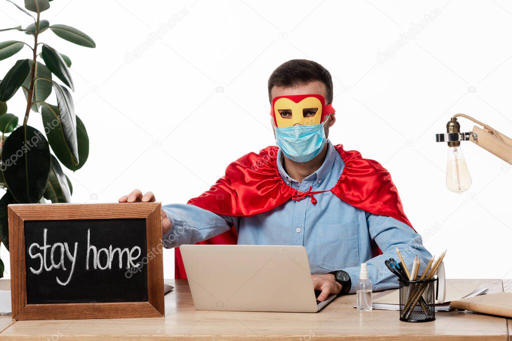 freelancer in medical mask and superhero costume holding chalk board with stay home lettering isolated on white 