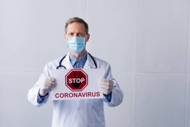 Beyaz önlüklü ve tıbbi maskeli orta yaşlı doktor elinde gri harfli Coronavirüs yazan bir levha tutuyor.
