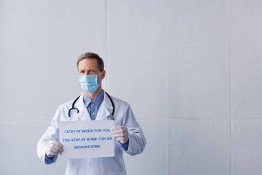Medikal maskeli orta yaşlı doktor pankart tutuyor. Ben senin için çalışıyorum, sen evde kalıp gri harfleri yazıyorsun.