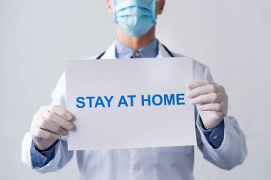 Orta yaşlı, maskeli, elinde plaketi olan, evde gri harflerle oturan bir doktor.