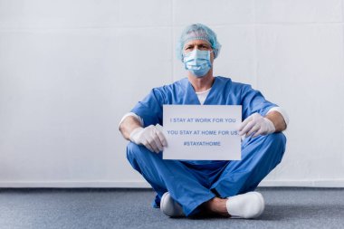Tıbbi maske takmış, şapka takmış olgun bir doktor. Ben senin için çalışıyorum, sen evde kalıp beyaz harflerle yazıyorsun. 