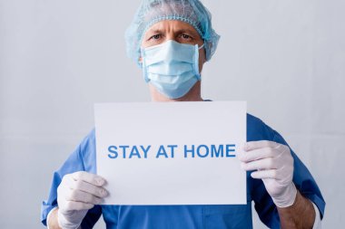 Medikal maskeli orta yaşlı bir doktor ve elinde gri harflerle evde duran bir pankart var.