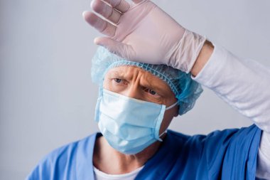 Mavi tıbbi maskeli ve şapkalı yorgun ve olgun bir doktor gri alnına dokunuyor. 