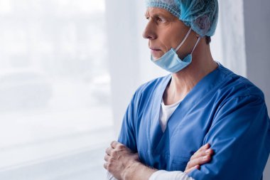 Mavi şapkalı ve tıbbi maskeli olgun bir doktor klinikteki pencereye bakıyor. 