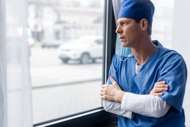 Mavi şapkalı ve tıbbi maskeli olgun bir doktor. Kollarını kavuşturup pencereye bakıyor. 
