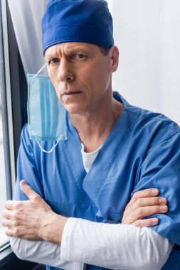 Mavi şapkalı ve tıbbi maskeli olgun bir doktor çapraz kollarla ayakta duruyor. 