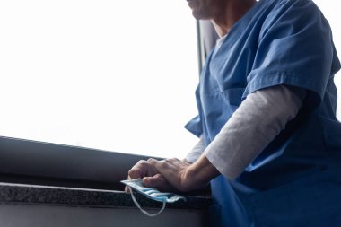 Tıbbi maske takan ve hastanenin penceresinin yanında duran olgun bir doktorun görüntüsü. 