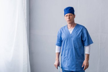Mavi şapkalı olgun bir doktor. Elinde tıbbi maske var ve gri üzerinde duruyor.