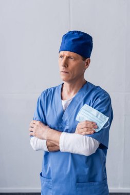 Orta yaşlı doktor mavi şapkalı çapraz kollu ve elinde gri maskeyle duruyor.