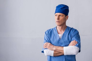 Mavi şapkalı düşünceli orta yaşlı doktor gri kollarıyla ayakta duruyor.