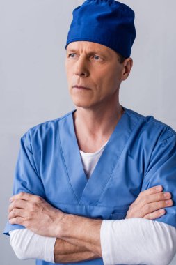 Mavi şapkalı düşünceli ve olgun doktor gri kollarıyla ayakta duruyor.