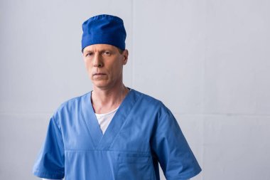 Mavi şapkalı olgun doktor gri kameraya bakıyor. 