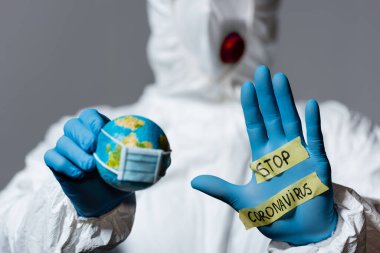 selective focus of man in hazmat suit holding globe and showing hand with stop coronavirus lettering isolated on grey  clipart