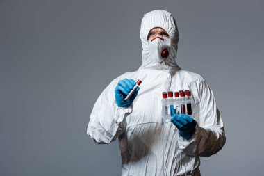 Tehlikeli madde giysisi giymiş olgun bir doktor, tıbbi maske ve gözlük takıyor. Elinde Coronavirus harfleri gri harflerle test tüpü tutuyor. 