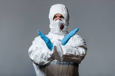 mature doctor in hazmat suit, medical mask and latex gloves standing with crossed arms isolated on grey  clipart