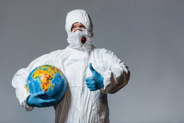 Tehlikeli madde giysisi ve tıbbi maskeli bir adam dünyayı tutuyor ve parmağının gri üzerinde olduğunu gösteriyor. 