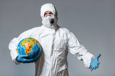 man in hazmat suit and medical mask holding globe and gesturing isolated on grey  clipart