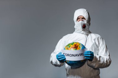 man in hazmat suit and medical mask holding globe and paper with coronavirus lettering isolated on grey  clipart