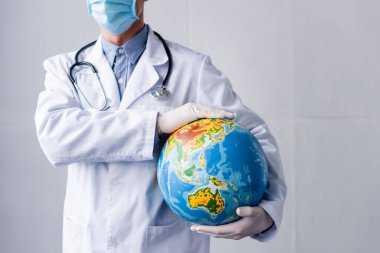 Lastik eldivenler ve gri üzerinde dünyayı tutan tıbbi maske takan olgun bir doktor görüntüsü. 