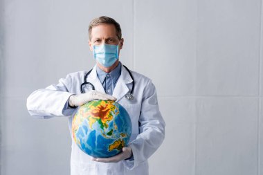 mature doctor in latex gloves and medical mask holding globe on grey  clipart