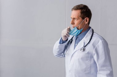 Tıbbi maskeli olgun bir doktor gri üzerine öksürüyor. 