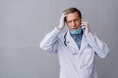 Stresli orta yaşlı doktor akıllı telefonla konuşuyor ve gri saçlara dokunuyor. 