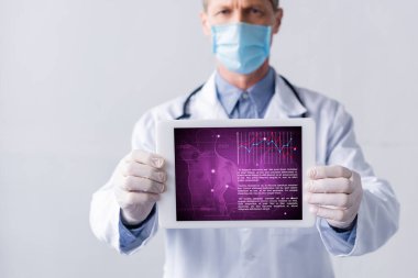 Tıbbi maskeli doktorun seçici odak noktası dijital tablet tutarken sağlık takip cihazı gri üzerine izole edilmiş.