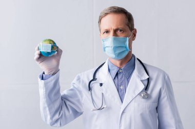 Lateks eldivenli ve tıbbi maskeli olgun bir doktor gri üzerinde küçük bir küre tutuyor.