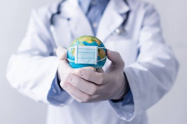 cropped view of middle aged doctor in latex gloves and white coat holding small globe in medical mask isolated on grey clipart