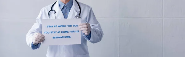 panoramic shot of middle aged doctor in latex gloves holding placard with i stay at work for you, you stay at home for us lettering on grey