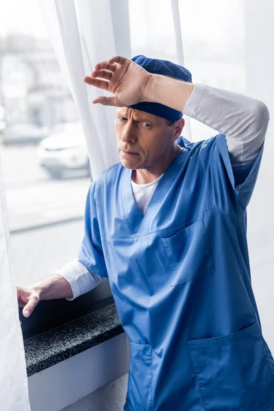 Exhausted Middle Aged Doctor Scrub Hat Touching Forehead Window Hospital — Stock Photo, Image