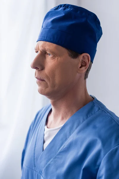 pensive middle aged doctor in scrub hat on white