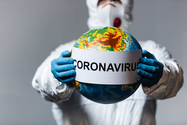 selective focus of man in hazmat suit and medical mask holding globe and paper with coronavirus lettering isolated on grey 
