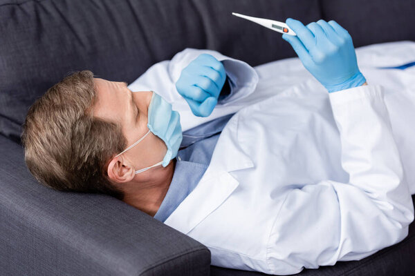 mature doctor in medical mask and latex gloves looking at digital thermometer while lying and coughing on sofa