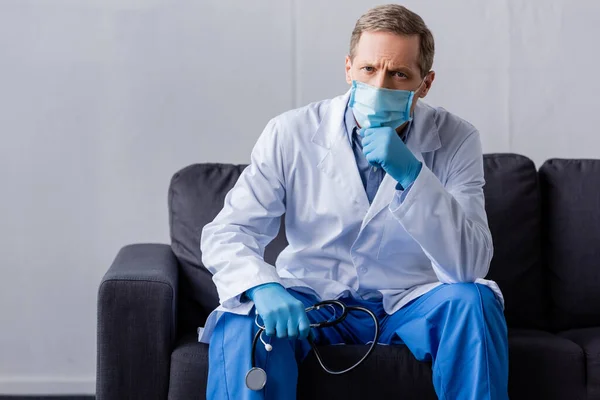 Dojrzały Lekarz Masce Medycznej Trzymając Stetoskop Siedząc Kanapie — Zdjęcie stockowe