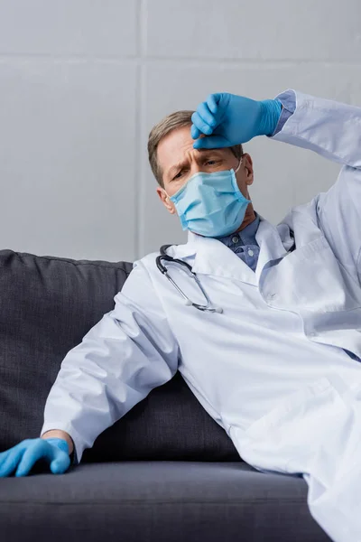 疲惫的成熟医生 戴着医用面罩和乳胶手套坐在沙发上 — 图库照片