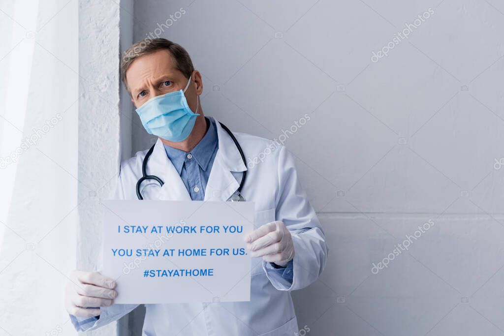 middle aged doctor in medical mask and latex gloves holding placard with i stay at work for you, you stay at home for us lettering on grey