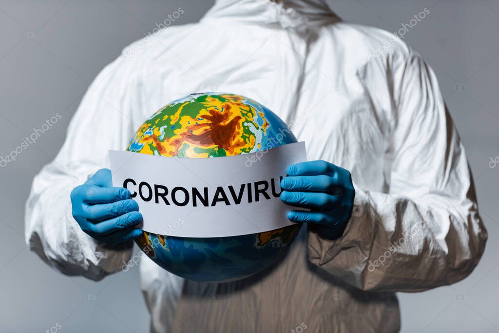 cropped view of man in hazmat suit holding globe and paper with coronavirus lettering isolated on grey 