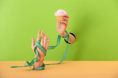 cropped view of woman with measuring tape on hands holding tasty cupcake on green and orange  clipart