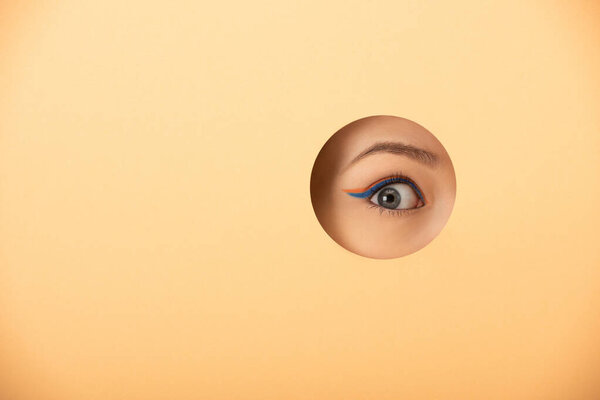 cropped view of woman with eye liner looking at camera through hole on pastel orange