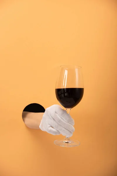 cropped view of woman in white glove holding glass of red wine on orange